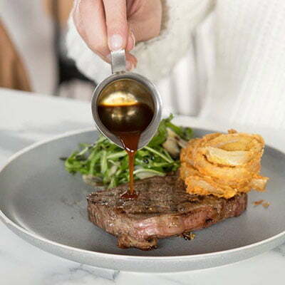 Person pouring gravy from a small jug onto a piece of steak.