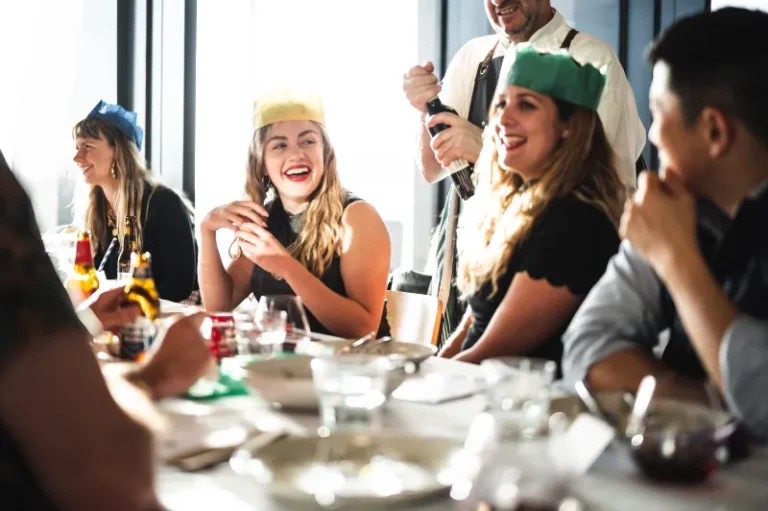 A group of people sitting at a restaurant table laughing while wearing coloured paper hats.