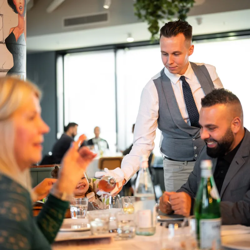 A waiter placing a plate of food on a table where two people look on happily.